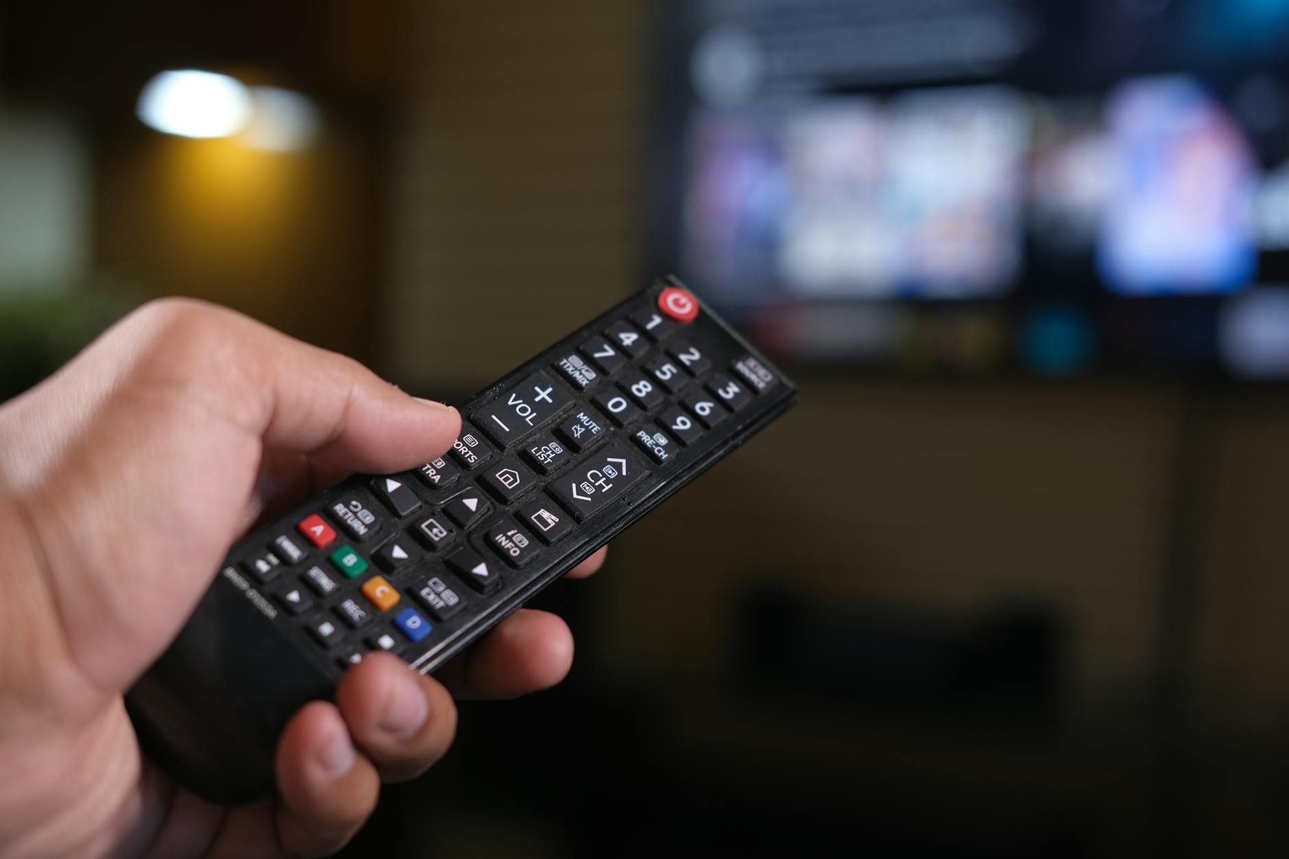 Hand holding TV remote control with multiple streaming service options displayed on screen